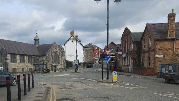 A picture showing the Lenten Pool junction in Denbigh. It is a 30way road junction with a mini roundabout in the middle. There are bus stops on two of the three sides. There is a church building visible (now run as the Body Studios gym), and a number of Victorian era brick buildings running up the hill from Bridge Street towards the High Street